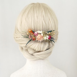 Puede incluir: Un peine para el cabello adornado con flores secas rosas, naranjas y blancas, con hojas verdes. El peine está colocado en cabello rubio peinado en un moño bajo. Un bonito accesorio para el cabello.