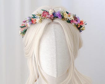 Könnte beinhalten: Ein farbenfroher Blumenkranz mit getrockneten Blumen in Rosa, Lila, Gelb und Grün. Der Kranz ist auf einem Schaufensterkopf mit langem, blondem Haar drapiert und erzeugt eine zarte und natürliche Ästhetik.