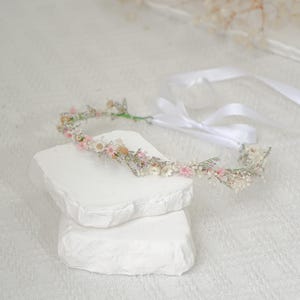 Peut inclure: Une couronne florale délicate réalisée avec des fleurs séchées dans des tons de blanc et de rose. La couronne est attachée avec un ruban de satin blanc et est présentée sur un socle en pierre blanche.