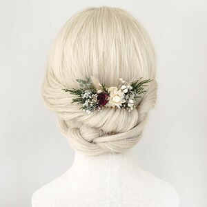 Puede incluir: Un peine para el cabello adornado con pequeñas flores secas en tonos burdeos, crema y blanco, acentuadas con follaje verde. El peine se coloca en cabello rubio peinado en un moño trenzado, para un look natural y elegante.