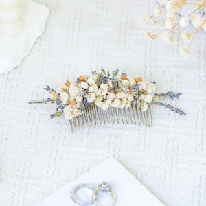 May include: A white and lavender dried flower hair comb. The comb is made of metal and has a delicate design. The flowers are arranged in a cluster and are accented with small, dried berries.