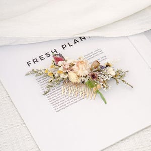 Puede incluir: Un peine para el cabello floral con una variedad de flores secas en tonos crema, rosa, amarillo y morado. El peine tiene dientes dorados y está sobre un fondo blanco con el texto "FRESH PLAN."