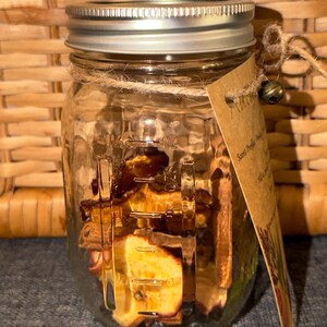 Puede incluir: Un tarro de cristal transparente lleno de frutas secas y especias. El tarro tiene una tapa plateada y una etiqueta de papel marrón atada con hilo. La etiqueta tiene texto. El tarro está sobre una superficie azul.