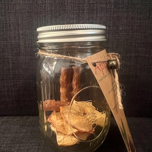 Puede incluir: Un tarro de cristal transparente con tapa plateada, lleno de rodajas de manzana deshidratadas y palitos de canela. Una etiqueta de papel marrón está atada al tarro con hilo. La etiqueta tiene la palabra "Apple" impresa.