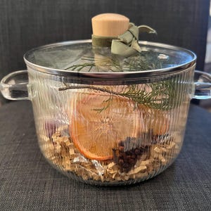 Puede incluir: Una olla de vidrio transparente con tapa, llena de rodajas de naranja secas, vegetación y especias. La olla tiene dos asas y un tapón de madera en la tapa, atado con una cinta verde.