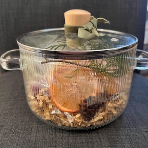 Può includere: Una pentola di vetro trasparente con coperchio, riempita con fette d'arancia essiccate, verdure e spezie. La pentola ha due manici e un pomello di legno sul coperchio, legato con un nastro verde. La pentola è su una superficie grigia.