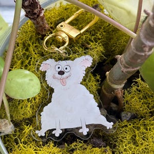 Può includere: Un portachiavi in acrilico trasparente con un cane dei cartoni animati con un corpo bianco e soffice, orecchie rosa e una lingua rosa. Il portachiavi ha una chiusura color oro. Il cane è seduto su muschio verde.