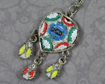 May include: A silver necklace with a teardrop-shaped pendant made of colorful mosaic tiles. The pendant has the word "ROMA" in the center and is surrounded by red, green, blue, and white tiles. The necklace has three smaller teardrop-shaped pendants hanging from the main pendant.