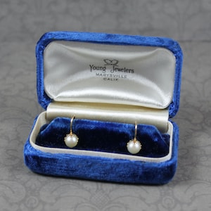 May include: Gold-coloured earrings with pearl drops presented in an open, blue velvet jewellery box. The box is lined with white satin and features the text "Young Jewelers, Marysville, Calif."