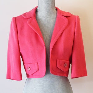 May include: A bright pink cropped blazer with short sleeves. The blazer has a notched collar, two front pockets with button closures, and a tailored fit. The jacket is displayed on a mannequin.