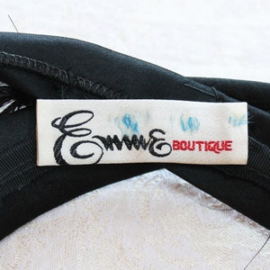 Puede incluir: Una diadema de tela negra con una etiqueta rectangular blanca cosida en el centro. La etiqueta muestra el texto en cursiva negra Emme y el texto en bloque rojo BOUTIQUE, con tenues marcas de tinta azul visibles sobre la tela blanca.