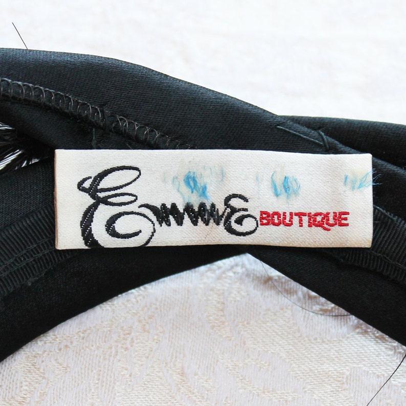 Puede incluir: Una diadema de tela negra con una etiqueta rectangular blanca cosida en el centro. La etiqueta muestra el texto en cursiva negra Emme y el texto en bloque rojo BOUTIQUE, con tenues marcas de tinta azul visibles sobre la tela blanca.