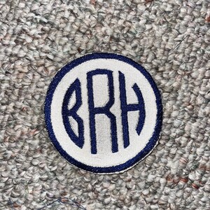 Puede incluir: Parche bordado circular con centro blanco y borde azul marino. El parche presenta las letras bordadas "BRH" en azul marino. El parche es probablemente para una chaqueta, bolso u otro artículo de tela.