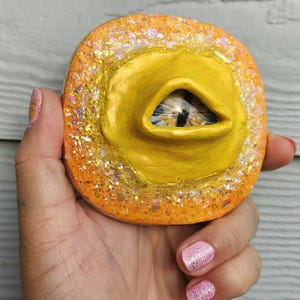 May include: A yellow and orange circular resin art piece with a glittery border and a yellow eye in the centre.