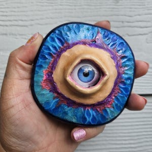 May include: A blue eye with a fleshy, textured, and colourful surrounding. The eye is set in a circular, fleshy, and textured frame. The frame is surrounded by a blue, purple, and pink textured background.