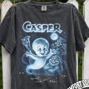 May include: A charcoal grey short-sleeve t-shirt featuring a graphic print of the character Casper the Friendly Ghost and three other ghosts above a city skyline. The word CASPER is printed in a glowing blue font above the characters.