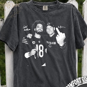 May include: A charcoal gray short-sleeve t-shirt featuring a black and white graphic of two individuals, one wearing a football jersey with the number 18. The graphic includes signatures and one person gesturing with a middle finger. The shirt has a crew neck.