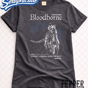 以下が含まれることがあります： 「Bloodborne」の文字とキャラクターのイラストが描かれた濃いグレーのTシャツ。「TONIGHT, GEHRMAN JOINS THE HUNT」の文字も含まれています。