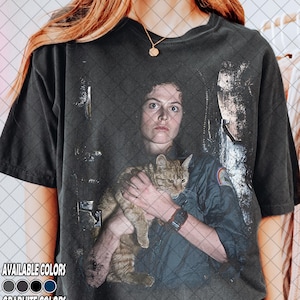 May include: Dark gray t-shirt featuring a distressed graphic of a woman holding a cat. The image includes the text "AVAILABLE COLORS" and "GRAPHITE COLORS" with color swatches. The shirt is a vintage style.