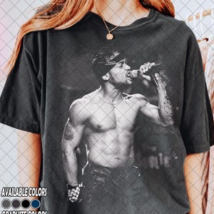 Può includere: T-shirt nero con grafica in bianco e nero di un uomo che canta in un microfono. L'immagine mostra l'uomo a torso nudo, con una bandana e gioielli. Il testo "AVAILABLE COLORS" e "GRAPHITE COLORS" è visibile.