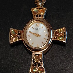 May include: A gold-tone Elgin pendant watch shaped like a cross, featuring an oval white face with gold-tone hands and numerals at 3, 6, 9, and 12. The cross arms are decorated with iridescent circular inlays and a gold-tone chain is attached.
