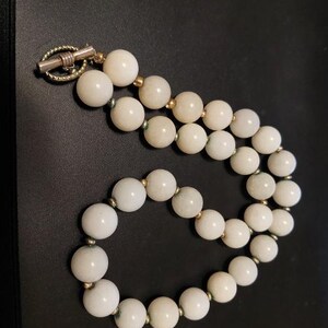 May include: A necklace composed of smooth, round, off-white beads separated by small metallic spacers. The piece features a silver-toned toggle clasp with a textured, twisted metal bar and a circular ring closure at one end of the strand.
