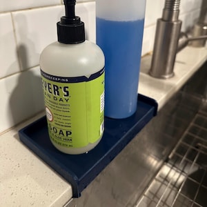 May include: A dark blue rectangular sink caddy tray designed to sit over the edge of a countertop. The tray holds a round bottle of Mrs. Meyer's Clean Day soap with a black pump dispenser and a tall, translucent blue bottle filled with liquid.