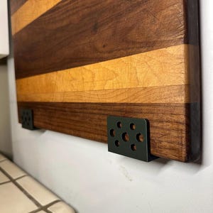 May include: A rectangular wooden cutting board featuring alternating horizontal stripes of dark brown walnut and light tan maple wood. The board is mounted to a wall using two black metal brackets, each with a pattern of six circular cutouts.