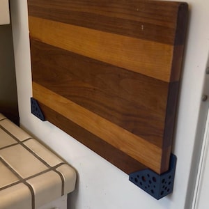 May include: A rectangular wooden cutting board with alternating dark and light brown stripes. The board is mounted on a white wall with two dark blue, right-angle brackets. The cutting board is a kitchen item.