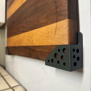 May include: A rectangular wooden cutting board with alternating dark brown and light tan horizontal stripes, secured to a vertical surface by two matte black metal corner brackets featuring a perforated circular pattern design.