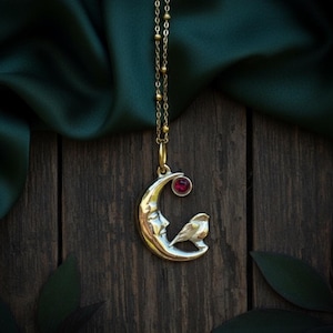 Może przedstawiać: Złoty naszyjnik z wisiorkiem w kształcie półksiężyca z twarzą, małym ptakiem i czerwonym kamieniem szlachetnym. Łańcuszek jest delikatny i złoty. Tło stanowi ciemna drewniana powierzchnia z zieloną tkaniną.