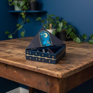 May include: A navy blue jewelry box with a celestial design. The box is open, revealing compartments for rings. The lid features a night sky scene with a moon, stars, and an owl. Gold star and diamond accents adorn the box.