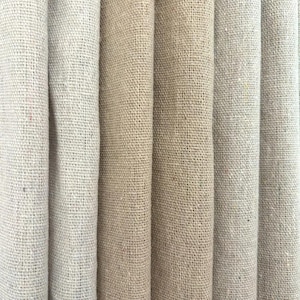 Puede incluir: Primer plano de varios rollos de tela de arpillera en diferentes tonos de beige y marrón claro. El tejido grueso de la tela es visible, creando una superficie texturizada. Los rollos están dispuestos verticalmente, mostrando los diferentes colores y texturas.