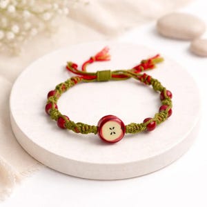 Op de afbeelding: Een handgemaakte armband met een rode appelvormige kraal met een wit kruisontwerp. De armband heeft rode kralen en is geregen op een groene gevlochten koord met rode en groene accenten. Gepresenteerd op een wit cirkelvormig oppervlak.