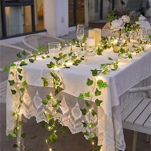 Puede incluir: Una mesa preparada para una reunión al aire libre, cubierta con un mantel blanco y decorada con guirnaldas de hiedra verde y pequeñas luces de hadas. Vasos transparentes y copas de champán están dispuestos sobre la mesa, junto con velas y faroles decorativos.