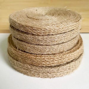 May include: A stack of coiled natural jute twine. The twine is a light brown color and appears to be tightly wound. The image is a close-up shot, highlighting the texture and construction of the twine.