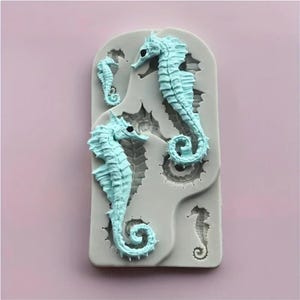 May include: A light blue silicone mould featuring three seahorse designs of varying sizes. The seahorses have detailed features and are set against a grey background. The mould is designed for crafting or baking, with a focus on marine-themed creations.