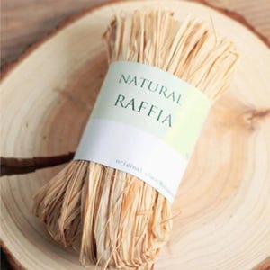 Può includere: Un fascio di rafia naturale legato con un'etichetta bianca e verde chiaro. L'etichetta recita "NATURAL RAFFIA" in un semplice font sans-serif. La rafia è di colore marrone chiaro e si trova su una superficie di legno.