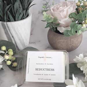 May include: A wax melt product in a clear plastic container, labelled "SEDUCTRESS." The label lists cocoa butter, vanilla, and jasmine. The product weighs 85.05g. The image includes decorative plants and flowers, creating a natural aesthetic.