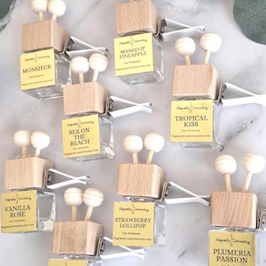 May include: A collection of small, square glass car air freshener bottles with light wood caps, two wooden diffuser sticks, and a white metal vent clip. Each bottle features a yellow label with scent names like Monsieur, Mango & Pineapple, Tropical Kiss, Sex on the Beach, Vanilla Rose, Strawberry Lollipop, Plumeria Passion, and Rose Petals.