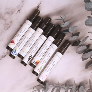 Puede incluir: Vista cenital de varios frascos roll-on de aceites esenciales con etiquetas blancas y tapas marr&oacute;n oscuro. Las etiquetas presentan ilustraciones y nombres de fragancias como "Vanilla Rose" y "Rose Petals". Los frascos est&aacute;n dispuestos sobre una superficie de m&aacute;rmol, con hojas de eucalipto.