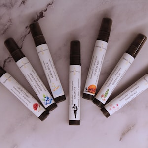 Puede incluir: Vista cenital de siete frascos de spray de aromaterapia marrones y blancos dispuestos en c&iacute;rculo. Cada frasco tiene una tapa negra y una etiqueta con texto e ilustraciones. Las etiquetas incluyen los nombres: Vanilla Rose, Blueberry Nuzzle, y Rose Petals. La botella central es Casanova.