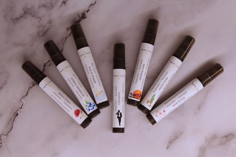 Puede incluir: Vista cenital de siete frascos de spray de aromaterapia marrones y blancos dispuestos en c&iacute;rculo. Cada frasco tiene una tapa negra y una etiqueta con texto e ilustraciones. Las etiquetas incluyen los nombres: Vanilla Rose, Blueberry Nuzzle, y Rose Petals. La botella central es Casanova.