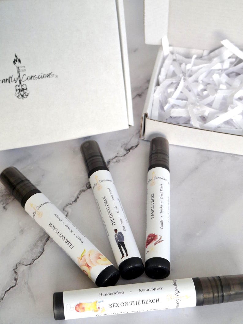 Puede incluir: Se muestran cuatro botellas de spray ambiental con tapas negras y etiquetas blancas. Las etiquetas dicen "Elegant Peach", "The Gentleman", "Vanilla Rose" y "Sex on the Beach". Una caja blanca y papel triturado est&aacute;n en el fondo.