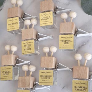 May include: A collection of car air fresheners with various scents. Each clear glass bottle has a wooden top with two spherical wooden diffusers. The labels display scent names like "Monsieur" and "Strawberry Lollipop".