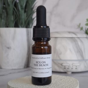 Puede incluir: Pequeña botella de vidrio ámbar con cuentagotas negro, etiquetada "SEX ON THE BEACH". La etiqueta enumera los ingredientes. La botella, que contiene 10 ml, está sobre una superficie blanca texturizada. Una planta y un difusor están en el fondo.