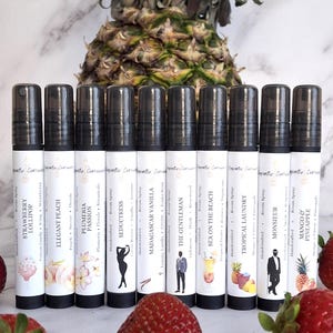 Puede incluir: Una colecci&oacute;n de aerosoles para habitaciones hechos a mano con tapas negras y etiquetas blancas. Cada etiqueta presenta un dise&ntilde;o &uacute;nico y un nombre de aroma, como "Strawberry Lollipop" y "Elegant Peach". Fresas y una pi&ntilde;a en el fondo.