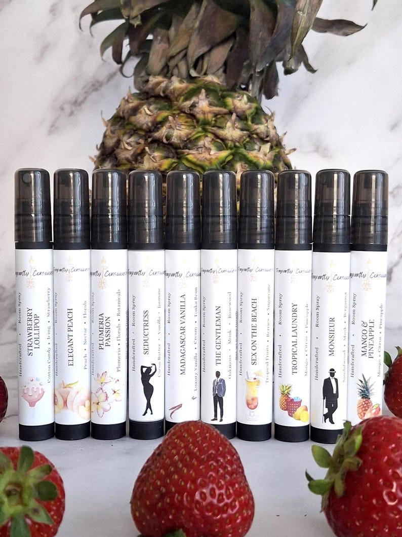Puede incluir: Una colecci&oacute;n de aerosoles para habitaciones hechos a mano con tapas negras y etiquetas blancas. Cada etiqueta presenta un dise&ntilde;o &uacute;nico y un nombre de aroma, como "Strawberry Lollipop" y "Elegant Peach". Fresas y una pi&ntilde;a en el fondo.