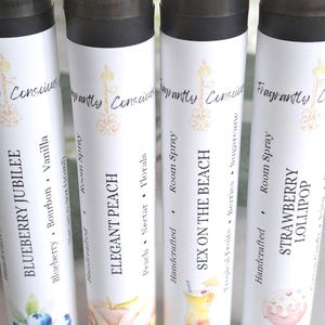 Puede incluir: Cuatro botellas de spray ambiental con tapas negras y etiquetas blancas. Las etiquetas presentan texto e ilustraciones para varios aromas: Blueberry Jubilee, Elegant Peach, Sex on the Beach y Strawberry Lollipop. Las botellas son artesanales.