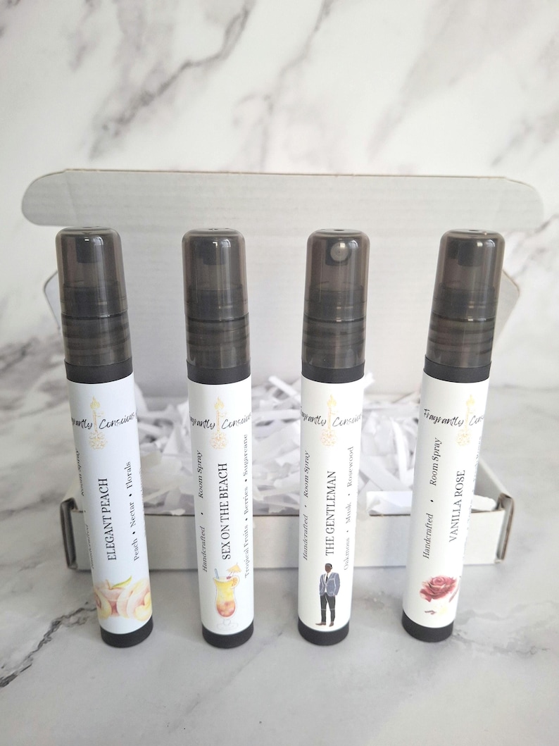 Puede incluir: Cuatro botellas de spray ambiental con tapas negras y etiquetas blancas dentro de una caja blanca. Las etiquetas dicen "Elegant Peach", "Sex on the Beach", "The Gentleman" y "Vanilla Rose". La caja est&aacute; sobre una superficie con vetas de m&aacute;rmol.